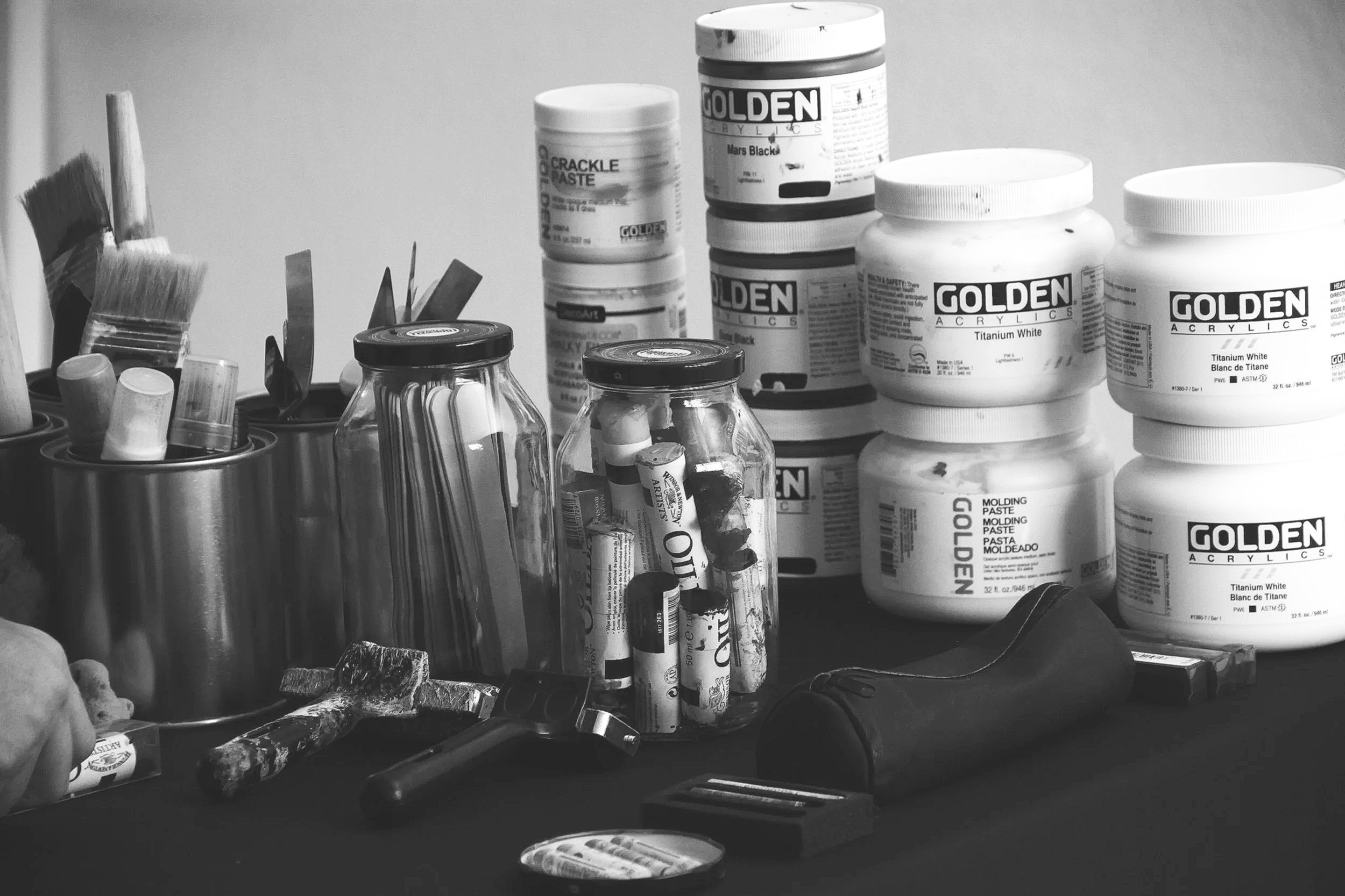 A black-and-white photograph documenting the painting materials and creative tools used by contemporary abstract artist Christopher Durst. Acrylic paints, brushes, oil sticks, texture mediums, and mixed media supplies are arranged within the artist's